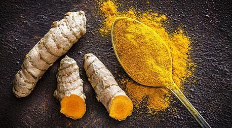 Fresh turmeric roots alongside a spoonful of bright yellow turmeric powder on a dark background.