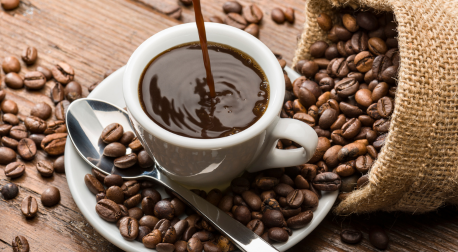 Hot coffee pouring into a white cup surrounded by roasted coffee beans on a wooden table.