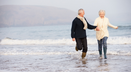 Active senior couple holding hands and running through beach waves, promoting a healthy lifestyle.