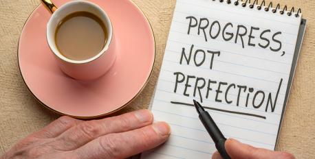 Handwritten note reading progress not perfection next to a pink coffee cup.