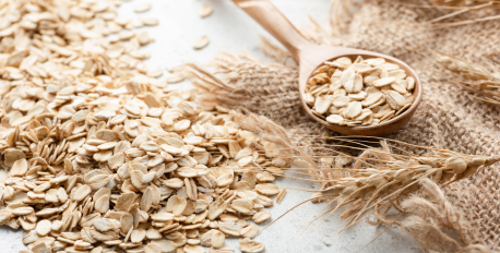 Rolled oats in a wooden spoon next to a pile of oats and wheat stalks on burlap.