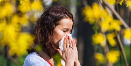 Woman sneezing into a tissue outdoors, surrounded by blooming yellow spring flowers.