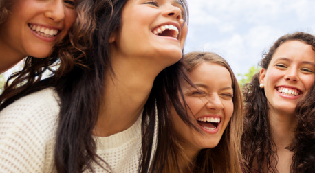 Four healthy, smiling young women laughing together outdoors under a bright sky.