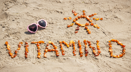 Vitamin D spelled with amber stones on sand next to a sun shape and pink sunglasses.