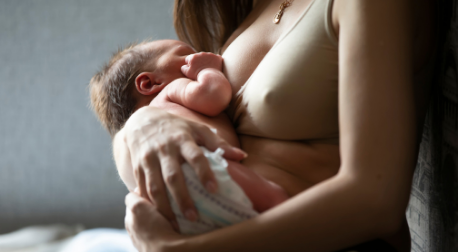 A mother in a beige nursing bra cradles and breastfeeds her newborn baby.