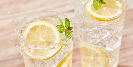 Two glasses of refreshing ice water with lemon slices and mint leaves splashing on a wooden table.