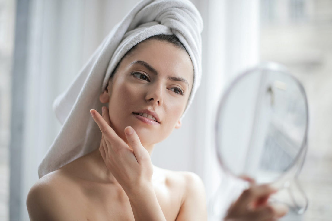 Woman with a white hair towel touching her face while looking in a hand mirror