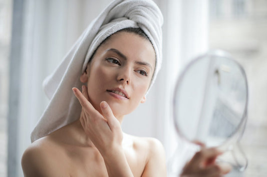 Woman with a white hair towel touching her face while looking in a hand mirror