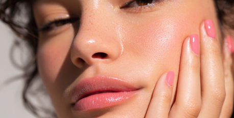 Close-up of a woman with glowing, hydrated skin and pink nails touching her cheek in natural sunlight