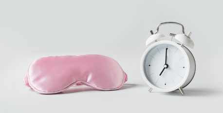 Pink silk sleep mask next to a white vintage alarm clock on a light background