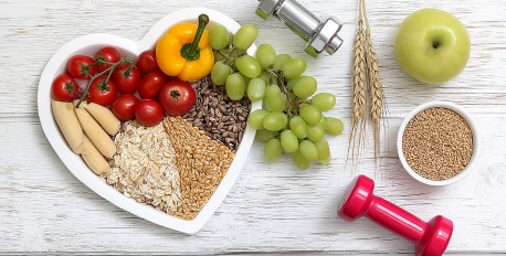 Heart-shaped bowl of healthy grains, fruits, and vegetables next to dumbbells on a white wooden background.