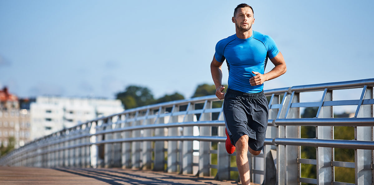 Man in a blue athletic shirt and dark shorts running across a bridge on a sunny day