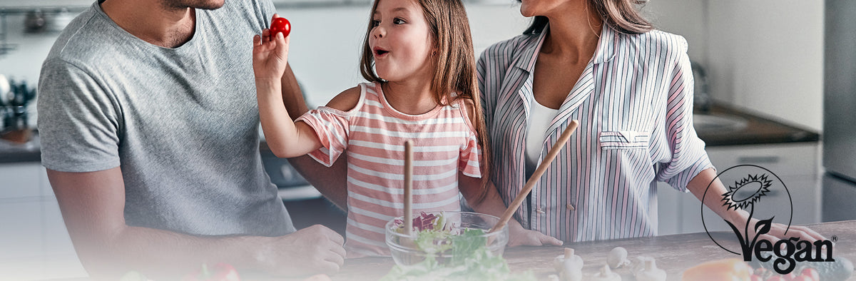 Family of three in a kitchen with a child holding a red object, surrounded by kitchen items.